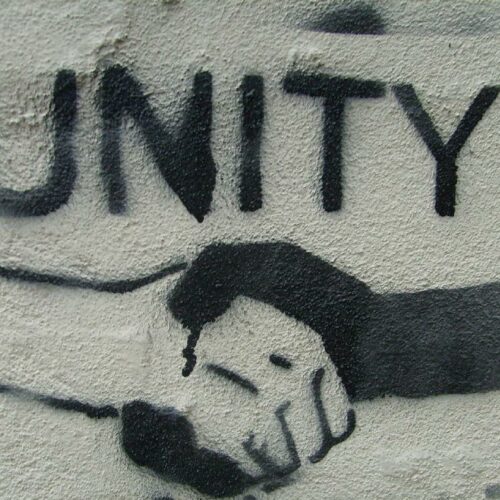 Ein Graffiti auf einer Hauswand, das zwei Hände zeigt, eine ist schwarz, die andere weiß. Darüber steht in schwarzen Großbuchstaben "Unity".
